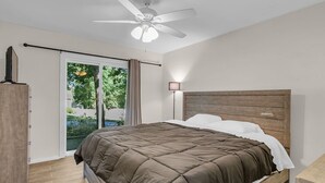 Un dormitorio con cama, ventilador de techo, mesita de noche, lámpara y vistas al exterior con vegetación.