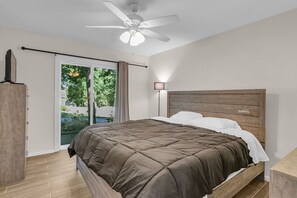 Ein Schlafzimmer mit Bett, Deckenventilator, Nachttisch, Lampe und Blick ins Grüne.