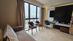 A modern hotel room with a large window, a sofa, two chairs, a desk with a TV, and a city view.