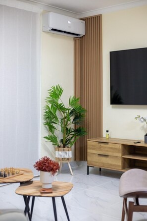 Ein modernes Zimmer mit einem Holztisch, einer Wand-Klimaanlage, einer großen Topfpflanze, einem Schachbrett und einem an der Wand befestigten Fernseher.