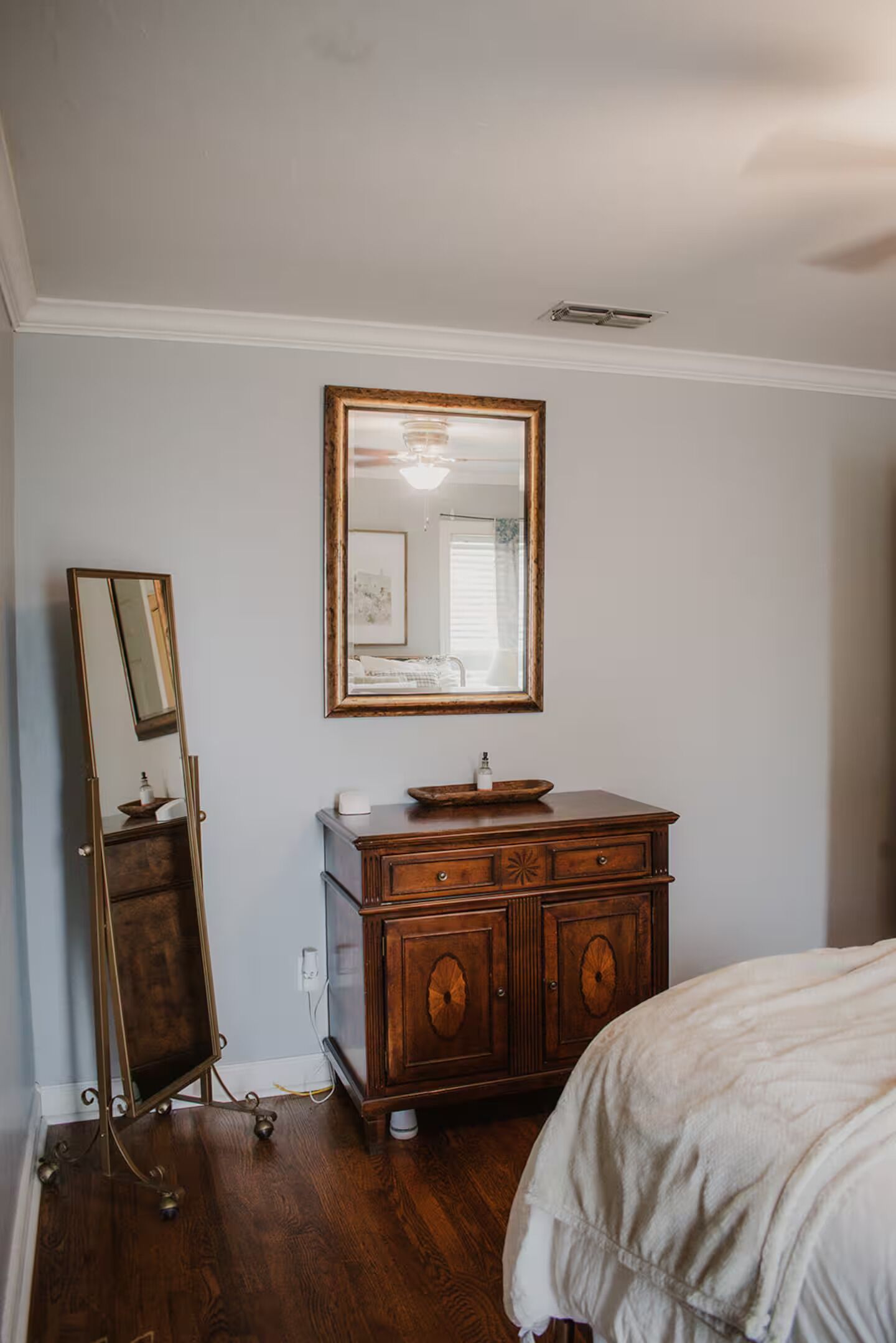 A bedroom with a wooden dresser, a full-length mirror, and a bed with a white comforter.