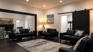Ein geräumiges Wohnzimmer mit dunklem Holzboden, zwei schwarzen Ledersofas mit gemusterten Kissen, einem runden Marmortisch, einer Stehlampe und einem großen Gemälde an der Wand.