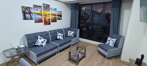 Una sala de estar con un sofá gris, una mesa de centro de vidrio y un ventanal con vista a un paisaje urbano.