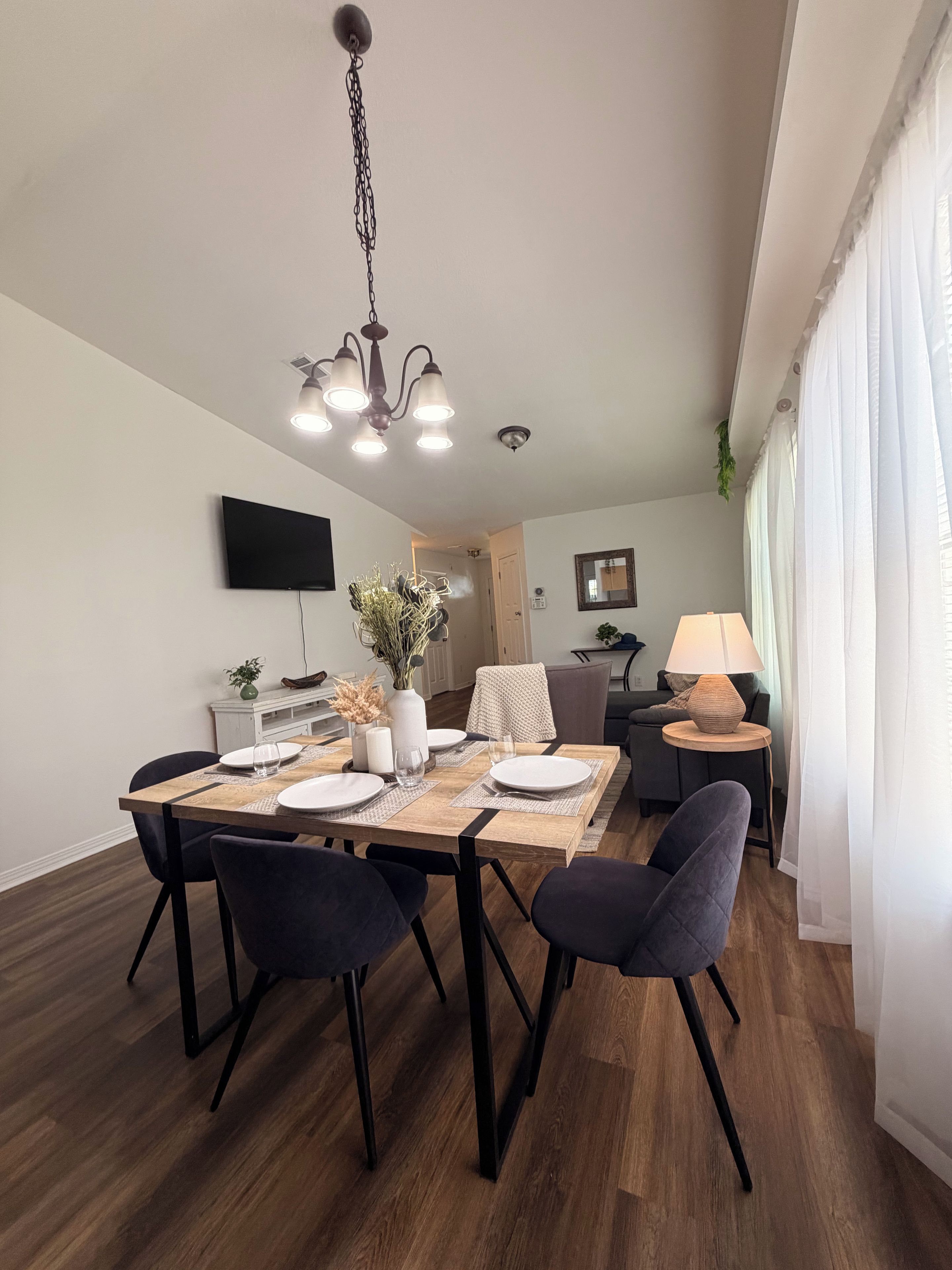 A dining area with a wooden table, black chairs, a chandelier, and a television on the wall.