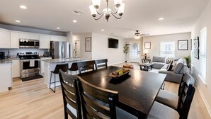 A modern kitchen with a dining area, a chandelier, and a living room with a sofa and a TV.