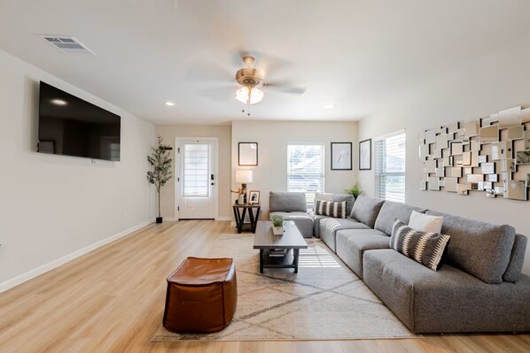 A modern living room with a sectional sofa, a coffee table, a brown ottoman, a ceiling fan, and a wall-mounted TV.