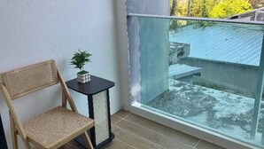 Ein Balkon mit Korbstuhl, einem kleinen Tisch mit Pflanze und Blick auf ein Schwimmbecken und Grünflächen.