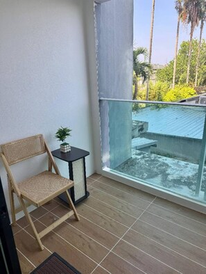 Ein Balkon mit Korbstuhl, einem kleinen Tisch mit Pflanze und Blick auf ein Schwimmbecken und Grünflächen.