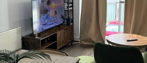 A living room with a TV, a wooden cabinet, a dining table with chairs, and a view of a red car outside.