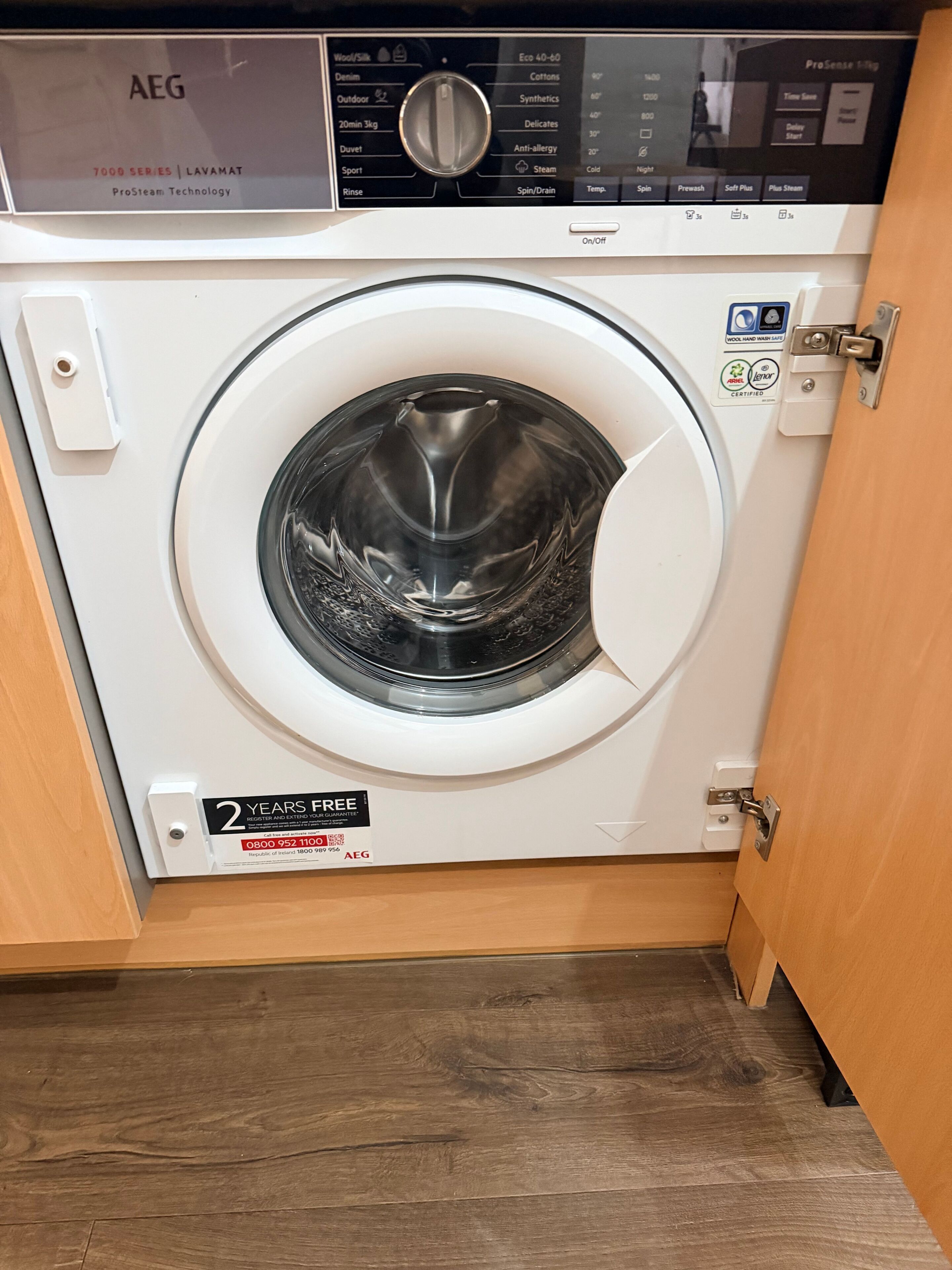 A front-loading washing machine with a digital control panel, a 2-year free registration offer, and a wooden floor.