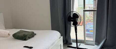 Una habitación de hotel con cama, ventilador, ventana con cortinas y vistas al exterior.