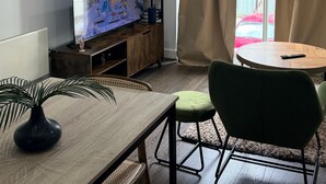 A modern living room with a flat-screen TV, a dining table with chairs, a small side table with a vase, and a window with curtains. - One Bedroom Flat in Mailbox With Free Parking (Birmingham)