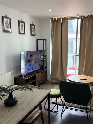 A modern living room with a flat-screen TV, a dining table with chairs, a bookshelf, and a large window with curtains. - One Bedroom Flat in Mailbox With Free Parking (Birmingham)