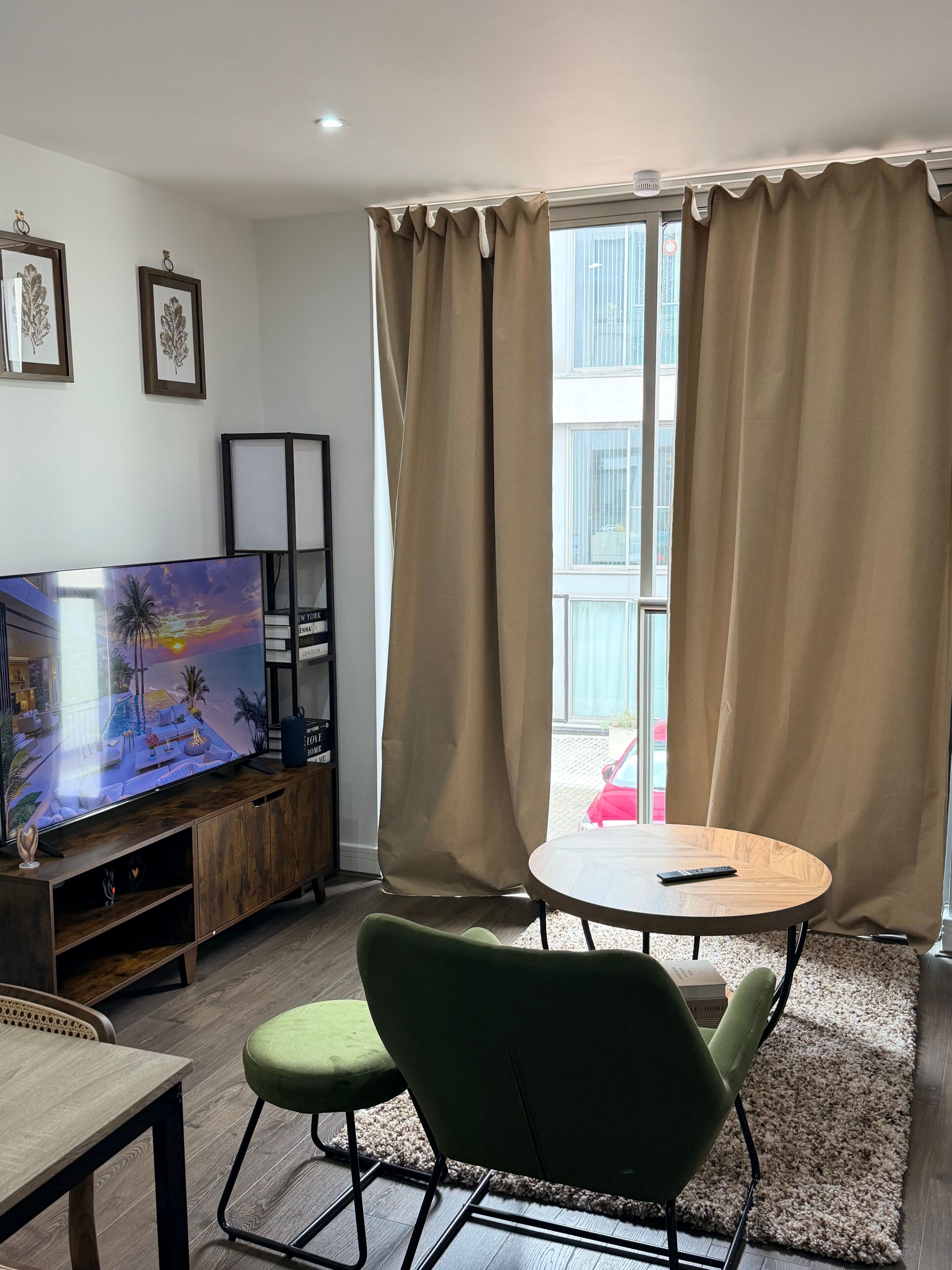 Un salon moderne avec un téléviseur à écran plat, une table ronde en bois, deux fauteuils verts, un meuble pour la télévision et une grande fenêtre avec des rideaux.