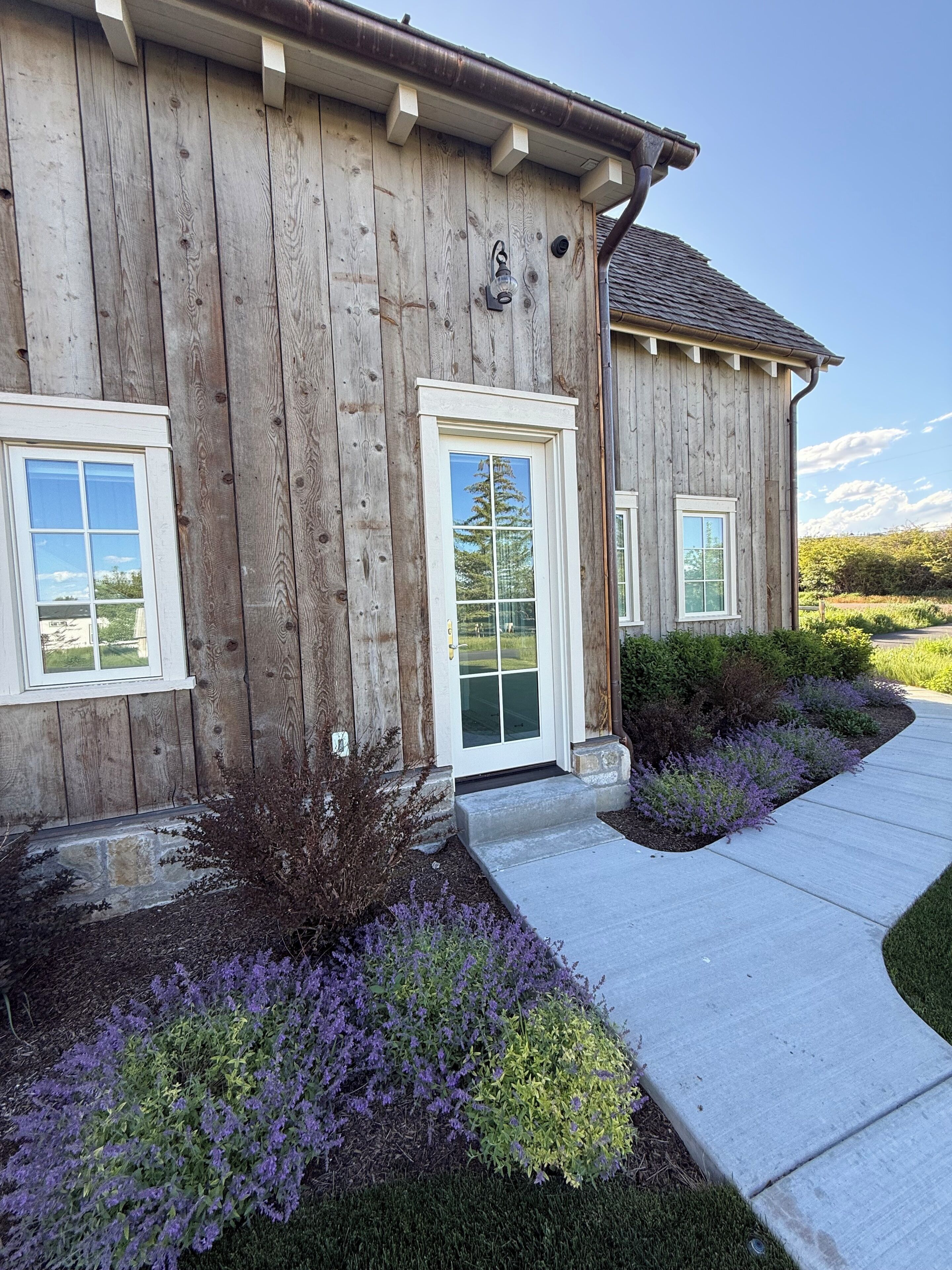 A wooden house with a front door, two windows, and a landscaped garden with purple flowers.