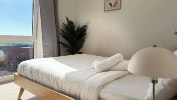 Une chambre avec une grande fenêtre, un lit recouvert de draps blancs, une plante en pot et une œuvre d’art accrochée au mur.