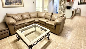 A living room with a sectional sofa, a glass-top coffee table, a framed picture on the wall, and a kitchen area in the background.