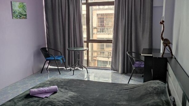 A bedroom with a bed, a small table, two chairs, a fan, and a view of buildings through a window. - Old Town (Pattaya)