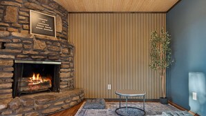 Un camino in pietra con un fuoco visibile, un pavimento in legno, un tappeto decorato, un piccolo tavolino rotondo e un pannello di parete in listelli di legno.