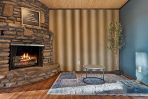 A stone fireplace with a visible fire, a wooden floor, a patterned rug, a small round table, and a vertical wooden slat wall. - Bella Vista MTB Escape • Large Patios Steps from Bike Trails (Bella Vista)