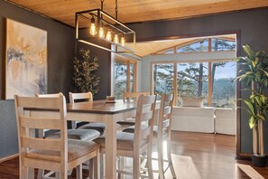 A dining area with a wooden table and chairs, a hanging light fixture, and a large window with a view of trees. - Bella Vista MTB Escape • Large Patios Steps from Bike Trails (Bella Vista)