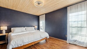 Una camera da letto con un letto in legno, un comodino, una lampada, una luce a soffitto e una finestra con tende leggere.