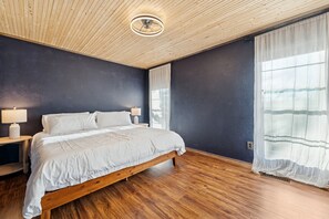 A bedroom with a wooden bed, a nightstand, a lamp, a ceiling light, and a window with sheer curtains. - Bella Vista MTB Escape • Large Patios Steps from Bike Trails (Bella Vista)