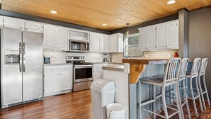 Una cucina moderna con mobili bianchi, elettrodomestici in acciaio inossidabile, soffitto in legno e sgabelli da bar.