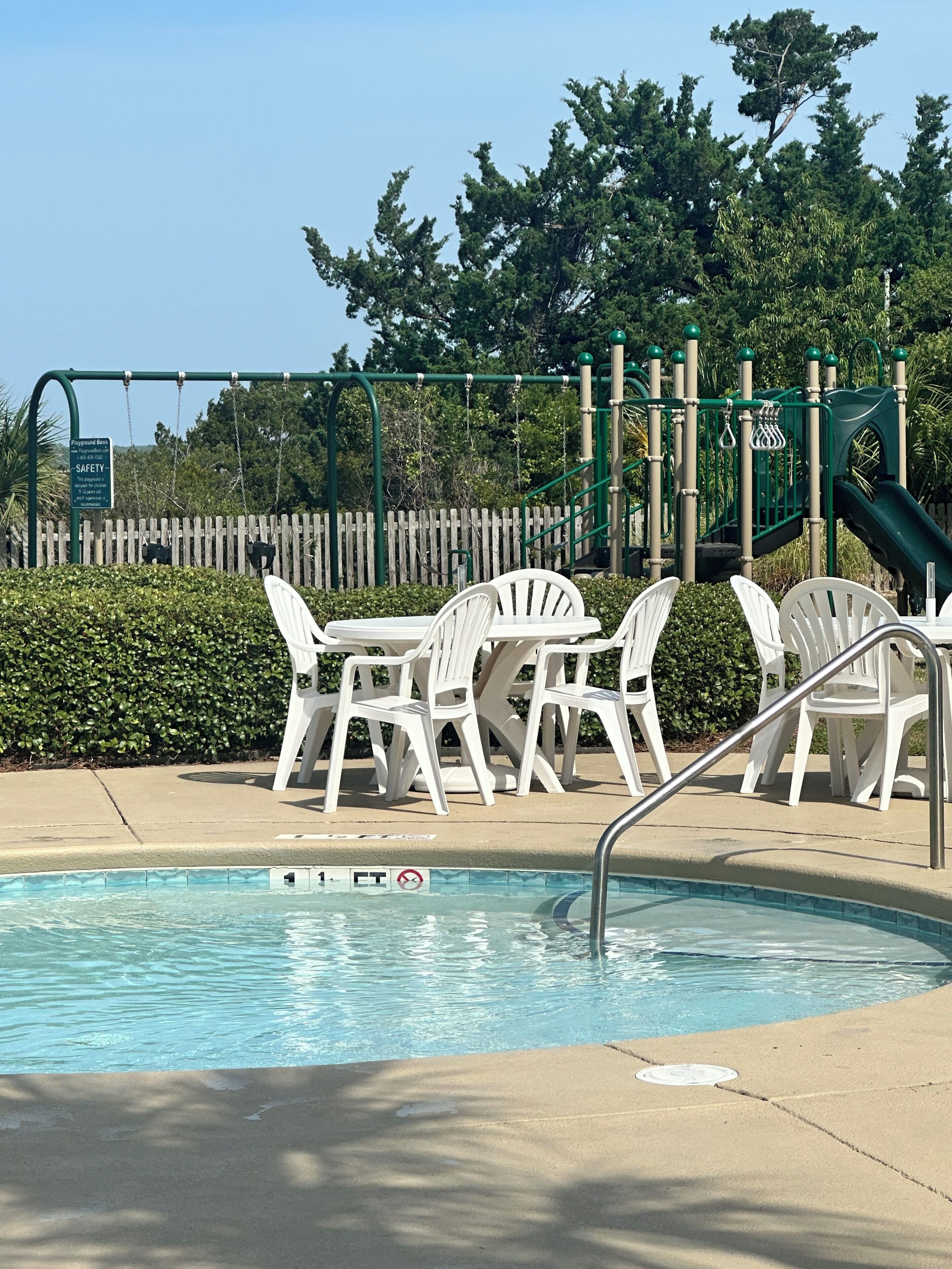 A swimming pool with a safety sign, white plastic chairs, and a playground structure in the background.