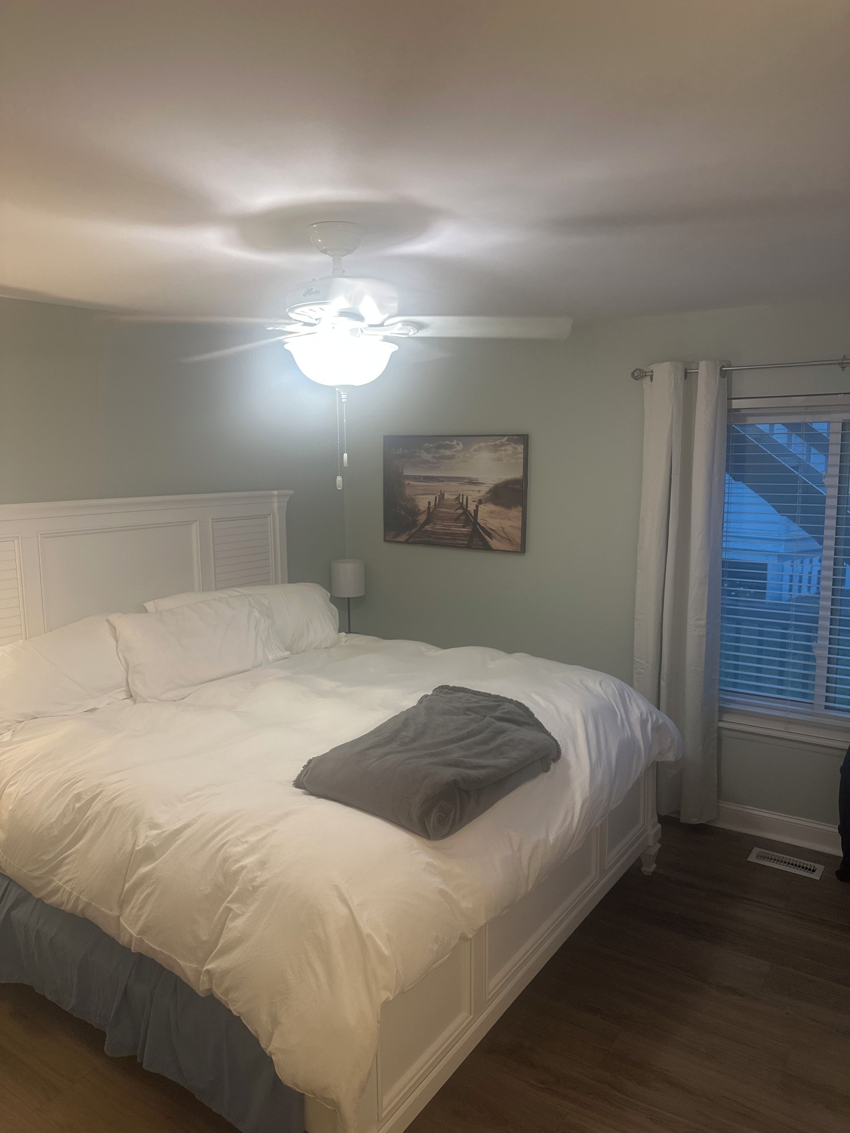 Ein Schlafzimmer mit Bett, Deckenventilator, einem Gemälde und einem Fenster mit Vorhängen.
