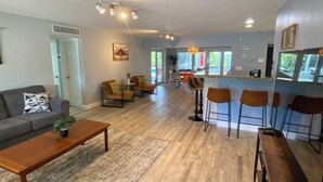 A modern living room with a sofa, coffee table, and bar stools. There is a kitchen area with a bar counter and a dining table. - Large Private Villa with Pool, Sauna, Billiards, Minutes from the Beach (Dania Beach)