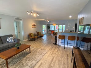 A modern living room with a sofa, coffee table, and bar stools. There is a kitchen area with a bar counter and a dining table. - Large Private Villa with Pool, Sauna, Billiards, Minutes from the Beach (Dania Beach)