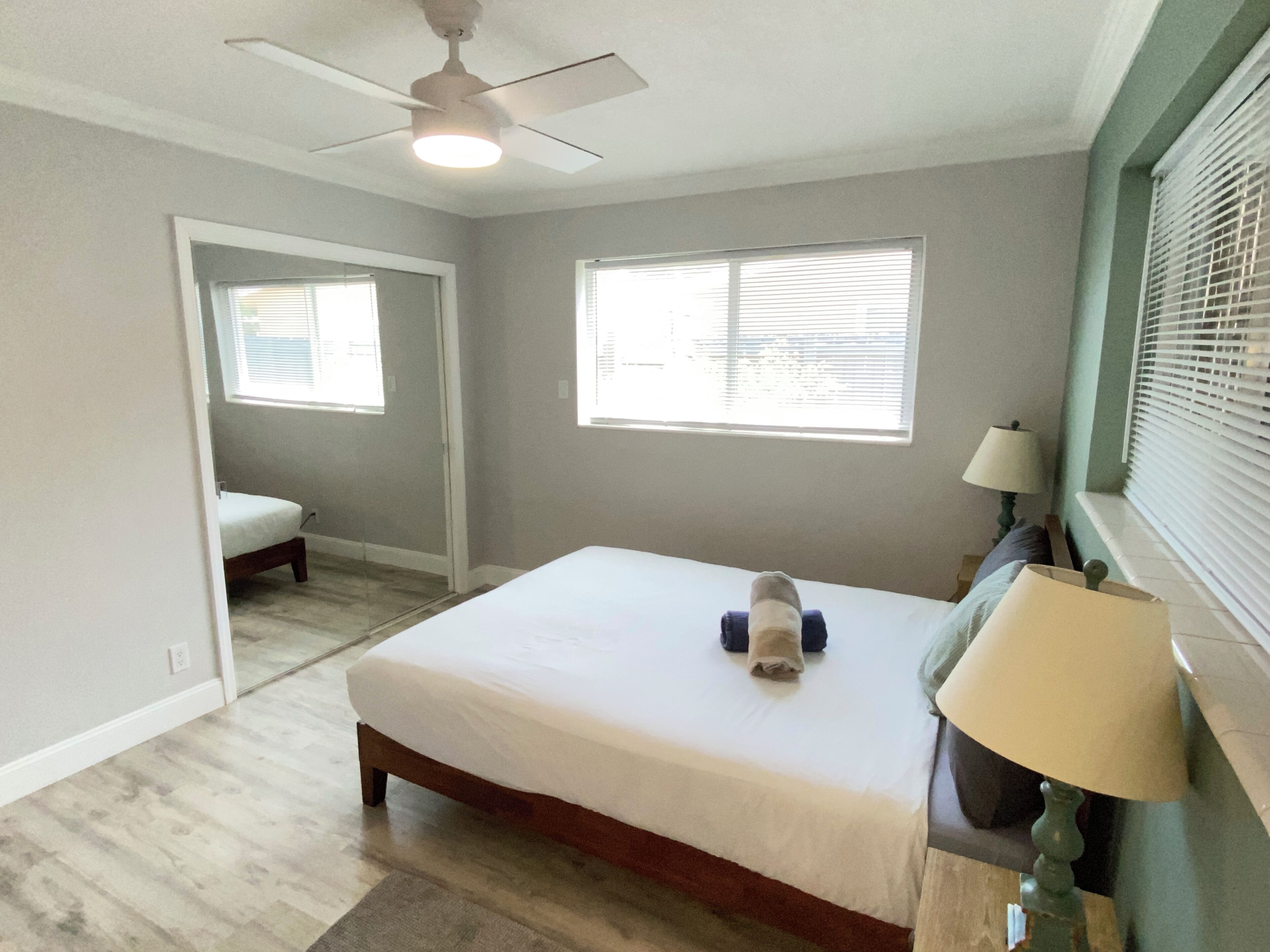 A bedroom with a bed, a ceiling fan, two windows with blinds, a lamp, and a door leading to another room.