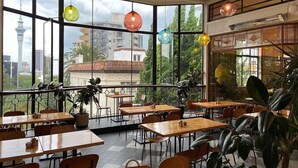 Ein Restaurant mit großen Fenstern, Holztischen und hängenden Pendelleuchten.