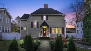 Ein zweistöckiges Haus mit Veranda, Schornstein und einem kleinen Vorgarten mit geschnittenen Büschen.