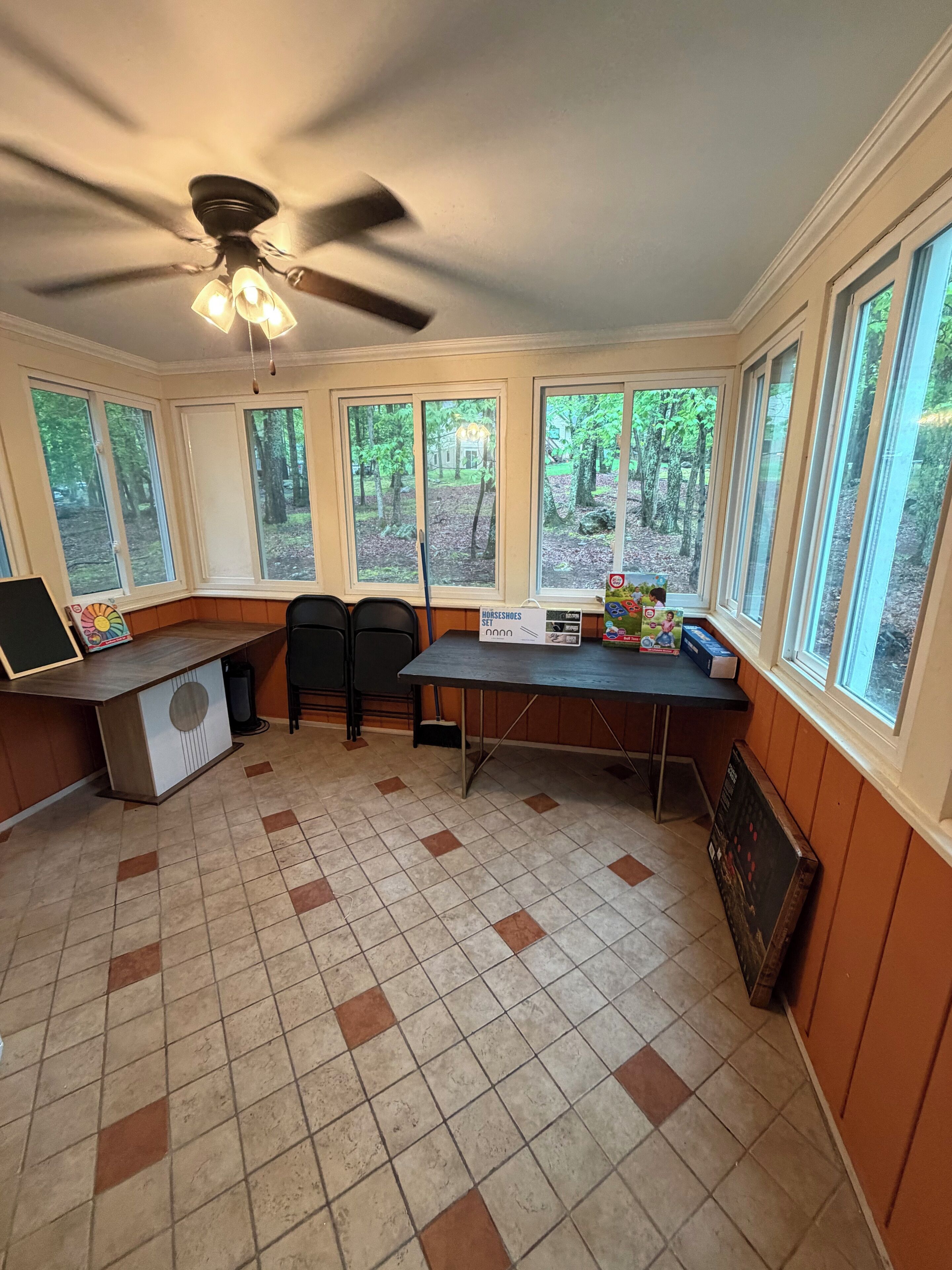 Habitación con escritorio, dos sillas, ventilador de techo y ventanas amplias con vistas a árboles.