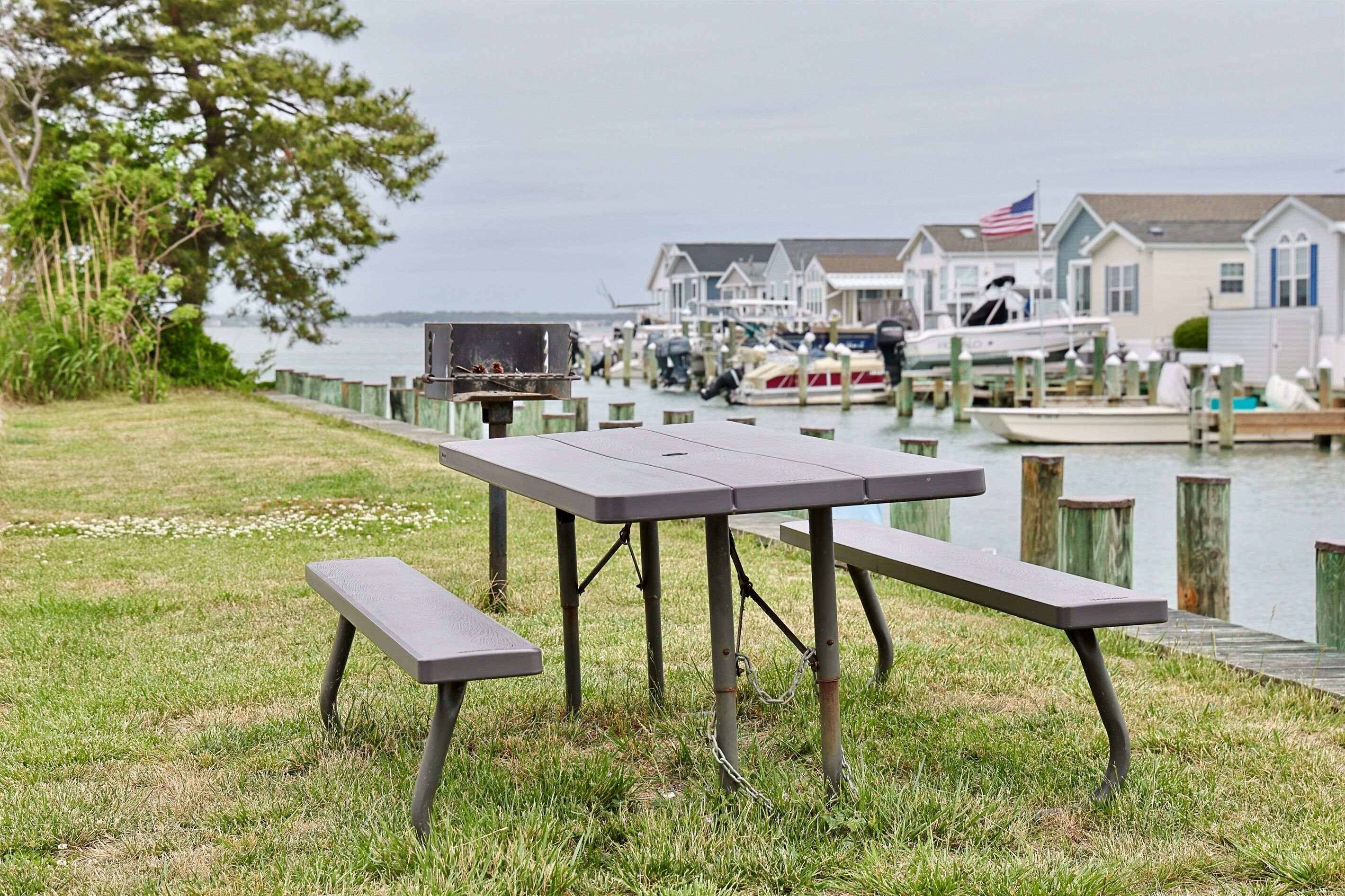 Une table de pique-nique et un banc sont installés au bord de l’eau, avec des maisons et des bateaux en arrière-plan.