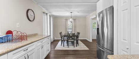 A kitchen with white cabinets, a stainless steel refrigerator, a dining table with chairs, and a clock on the wall.