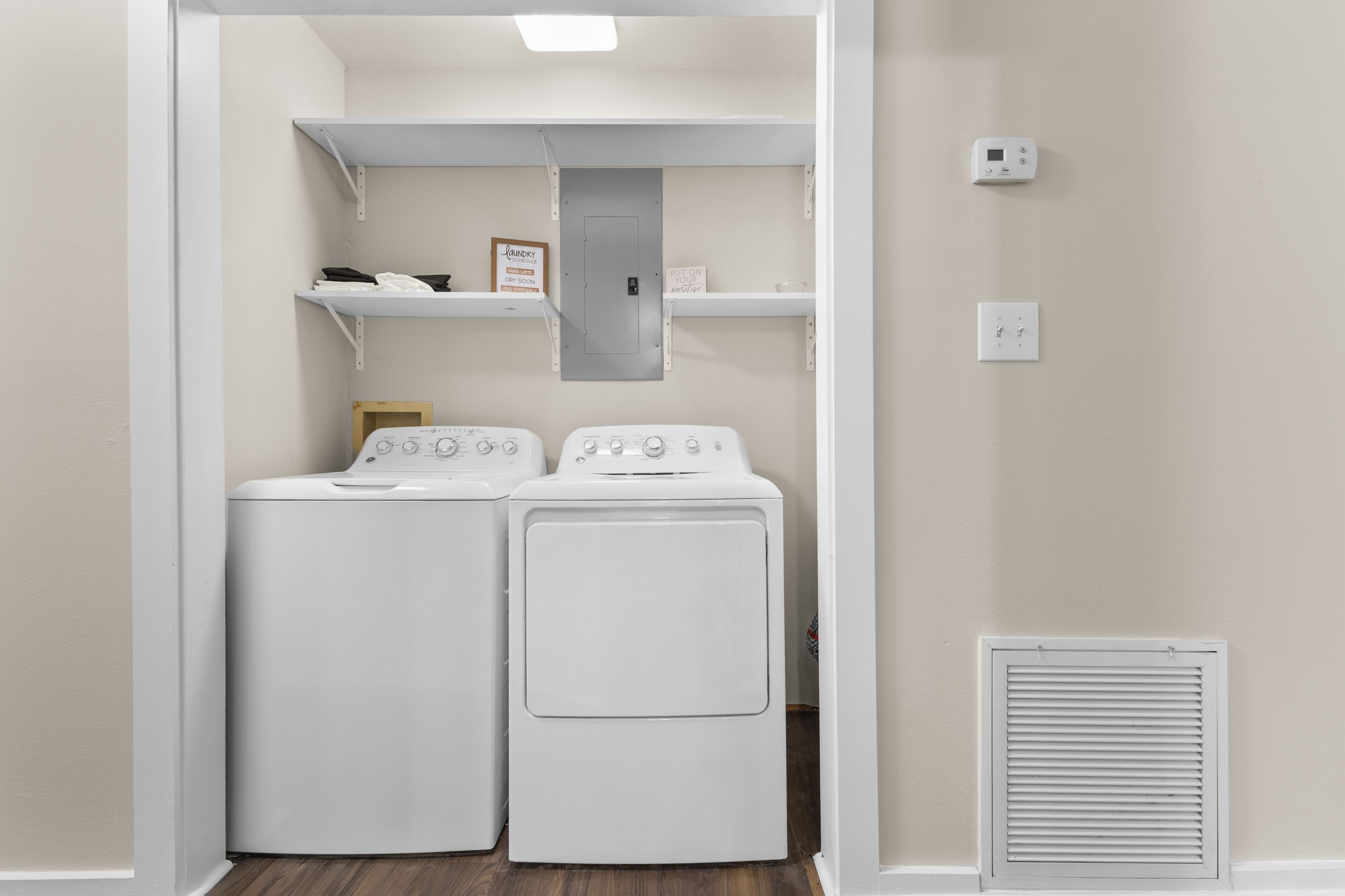 Une buanderie équipée de deux machines à laver et de deux sèche-linge à chargement frontal, tous deux de couleur blanche, d’une petite étagère sur laquelle est posée une photo encadrée, et d’une bouche d’aération au mur.