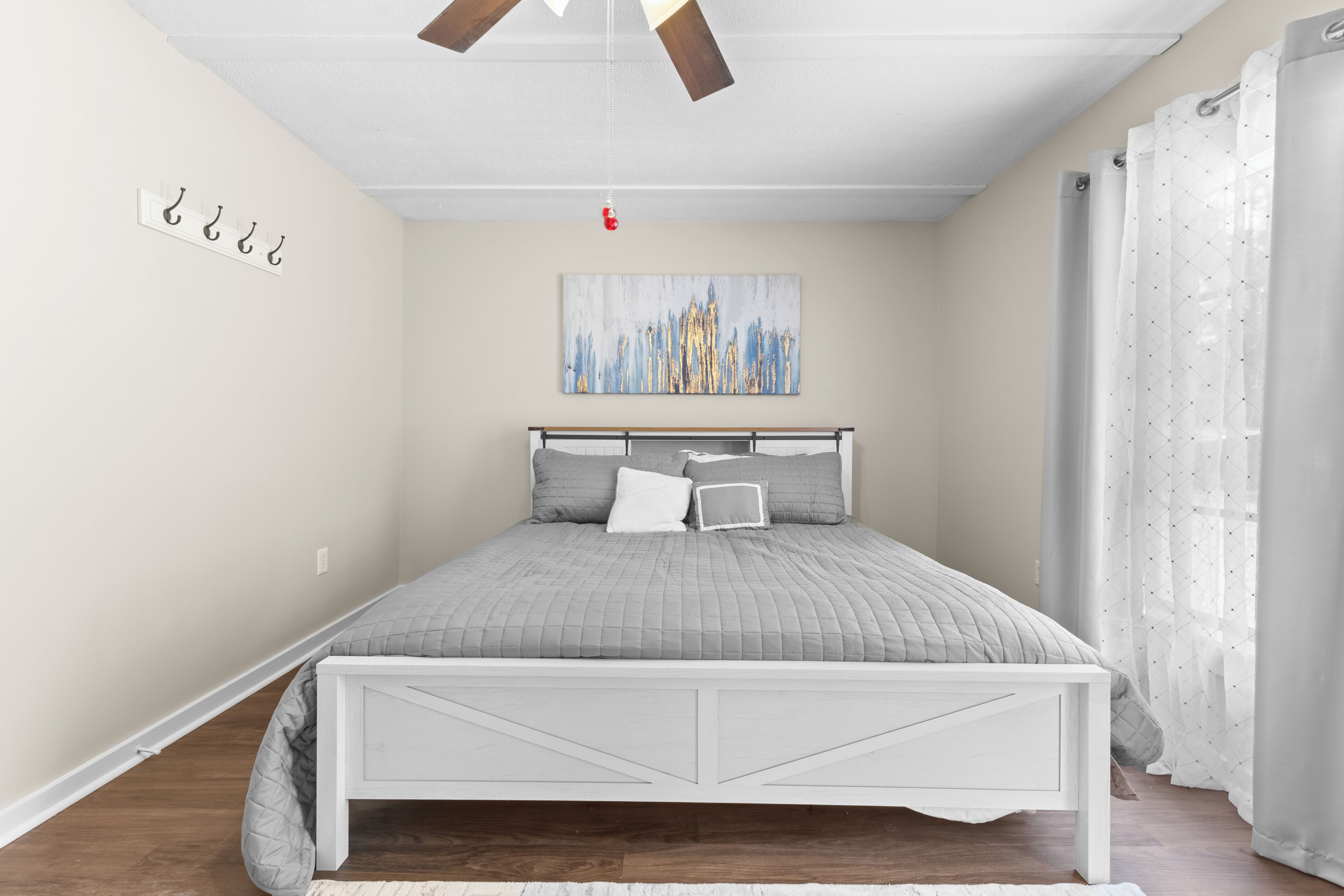 A bedroom with a bed, a ceiling fan, a wall-mounted light, a painting, and a window with curtains.