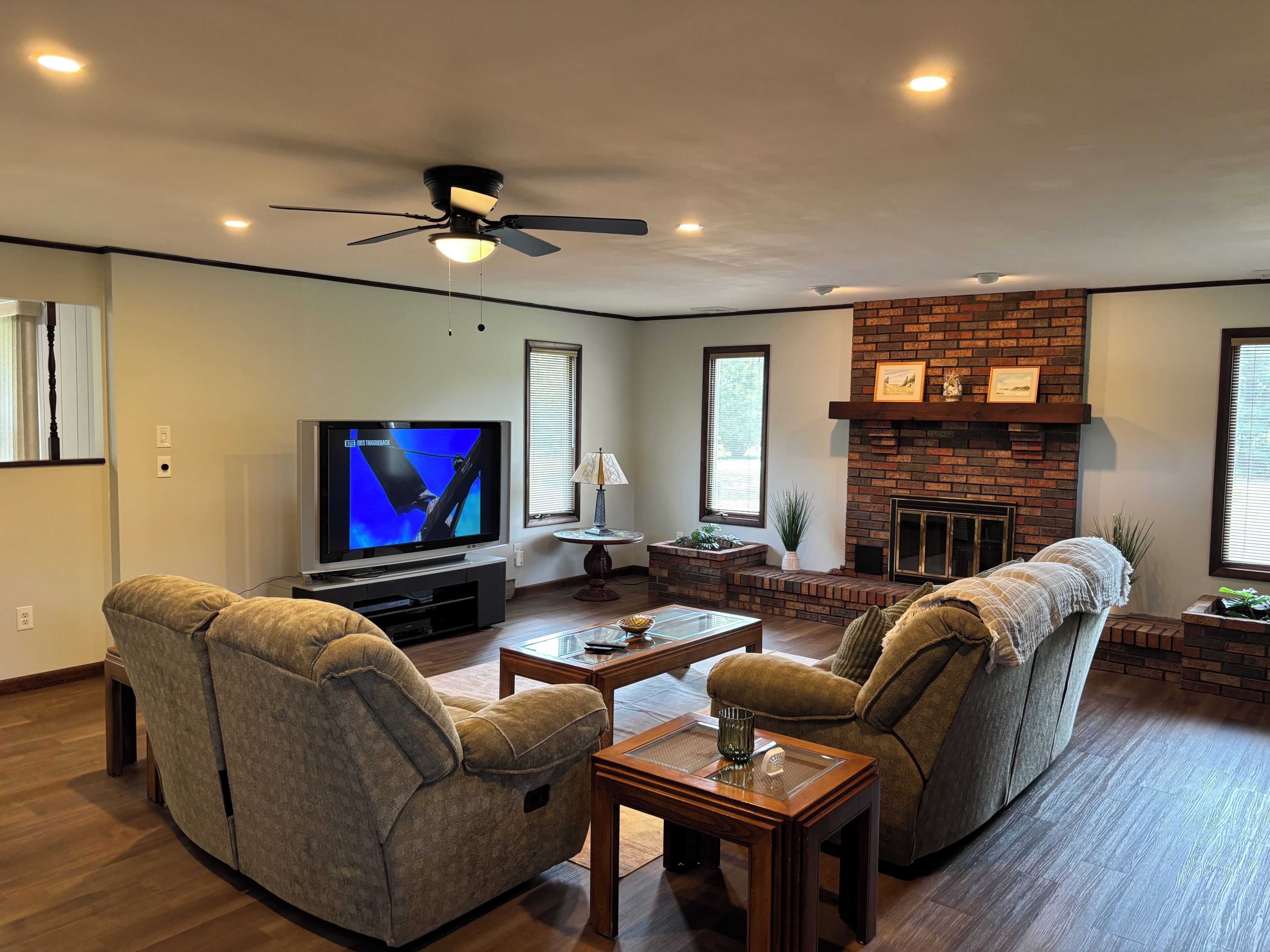 A living room with a flat-screen TV, two armchairs, a coffee table, a fireplace, and a ceiling fan.
