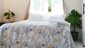 Ein Bett mit einer Bettdecke mit floralem und verschnörkelten Muster, eine Topfpflanze und ein Fenster mit Vorhängen.