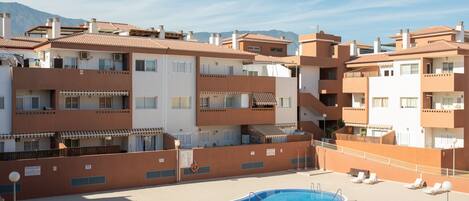 A rooftop swimming pool with lounge chairs, surrounded by multi-story buildings with balconies and a mountainous backdrop.