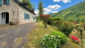 Una casa de piedra con camino de grava, rodeada de exuberante vegetación y montañas al fondo.