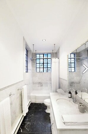 Un baño moderno con bañera, inodoro y zona de ducha, con paredes de mármol y suelo de baldosas negras.