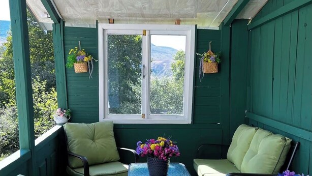 En inglasad veranda med gröna väggar, ett fönster med utsikt över grönskan och två stolar med kuddar.