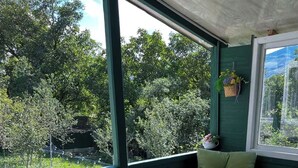 En inglasad veranda med ett bord och stolar, en hängande blomkorg och utsikt över träd och en byggnad.