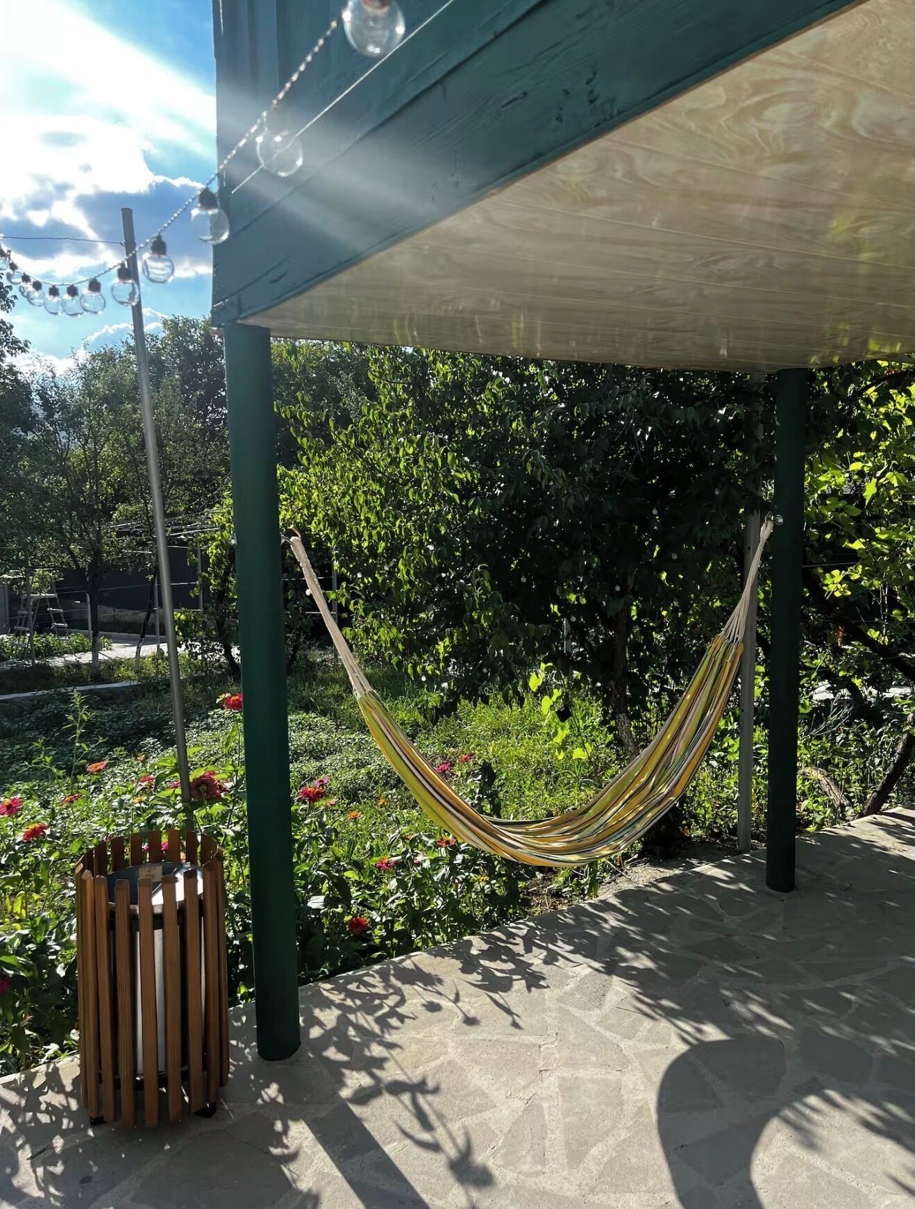 A hammock under a shaded structure in a garden with a trash bin nearby.
