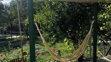 A hammock under a shaded structure in a garden with a trash bin nearby.