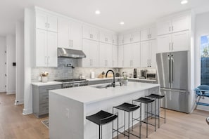 A modern kitchen with white cabinets, a stainless steel refrigerator, and a central island with bar stools. - Pool + Sauna | 2 Homes for Groups | Sleeps 14 | (Austin)